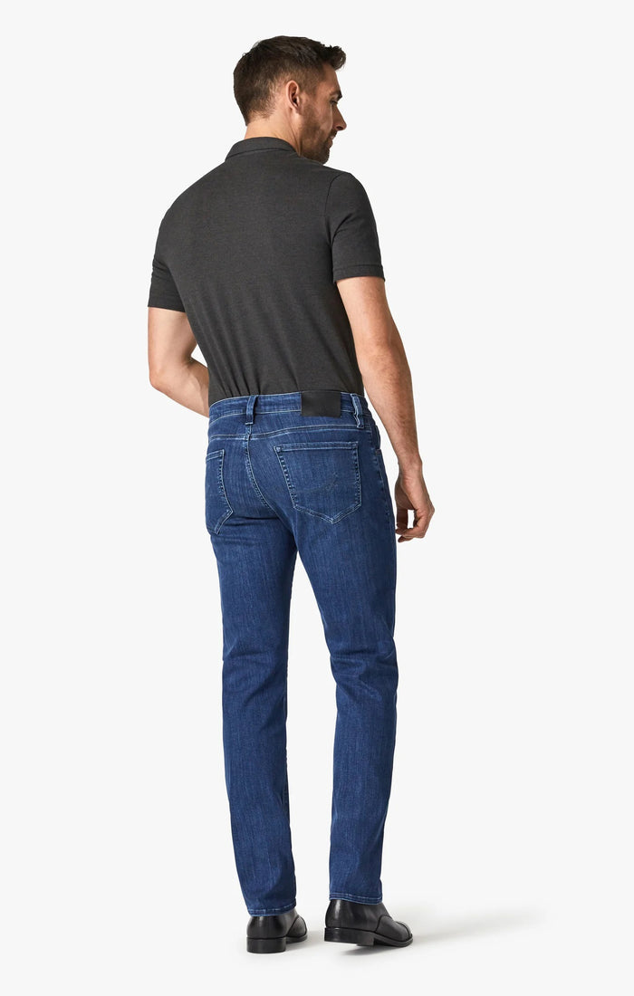 A man stands facing forward, wearing a dark short-sleeve polo, 34 HERITAGE - COURAGE STRAIGHT LEG JEAN IN MID URBAN WASH, a black belt, and black shoes. One hand is in his pocket; he has short hair, a trimmed beard, and a neutral expression.