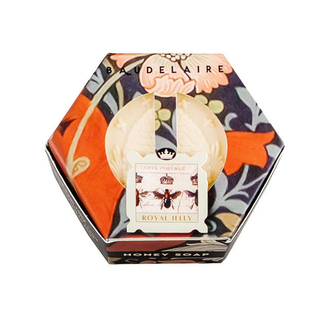 The BAUDELAIRE - ROYAL JELLY HONEY SOAP HEX BOX 3.5 OZ features a hexagonal box adorned with a floral design and "Baudelaire" elegantly written on top. At the center, a circular label showcases a vintage bee illustration with the text "Royal Jelly Soap." Inside, you’ll find honey-colored soap with an embossed design that promises to hydrate your skin for a glowing complexion.