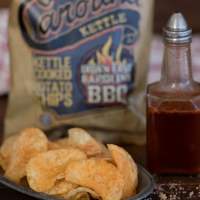 A bag of CAROLINA KETTLE - DOWN EAST BBQ CHIPS from 1 IN 6 SNACKS, featuring a brown and orange design with BBQ flames and bold text, delivers the authentic taste of Eastern North Carolina BBQ sauce accented by a hint of smoky charcoal flavor—making it a true standout among snack foods.