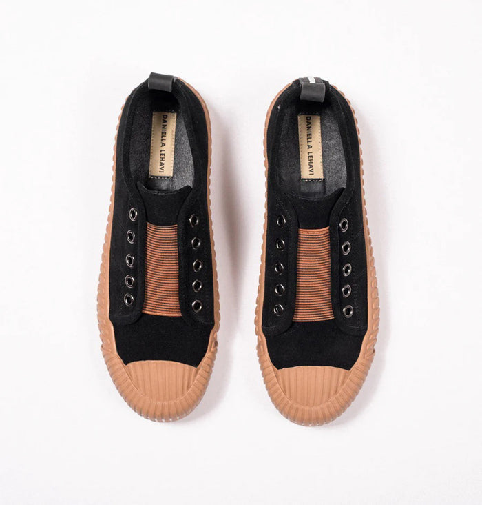 A pair of black slip-on sneakers with tan rubber soles and toe caps, viewed from above. The shoes have elastic bands where the laces would be and a black pull tab at the heel.