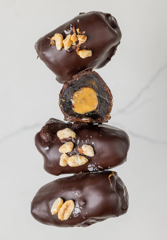 A white "DATE BETTER - PEANUT BUTTER CRUNCH COVERED DATES" package from DATE BETTER SNACKS rests on marble, cacao nibs and nuts spilling out. Orange text and illustrations adorn the packaging, adding charm to the scene.