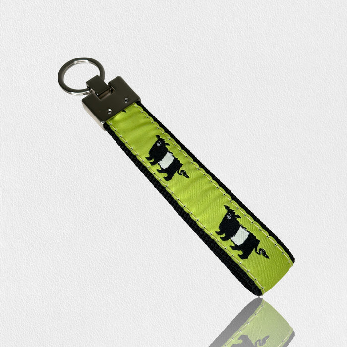 The UP COUNTRY FEARRINGTON SIGNATURE COLLECTION - BESPOKE BELTIE KEY CHAIN features green fabric, black edges, a metal ring, and a repeating black-and-white Beltie bear pattern—ideal for Fearrington fans. Shown on a white textured background.
