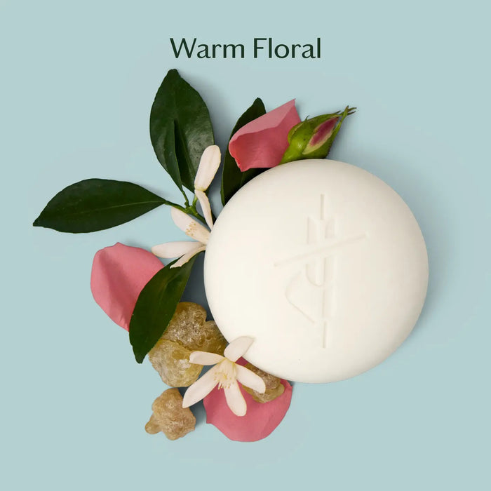 A round white KATE MCLEOD - DAILY STONE MINI LOTION BAR 1OZ, partially wrapped in beige cloth, rests beside its green box on a pale blue background. Crafted with natural essential oils for rich hydration.