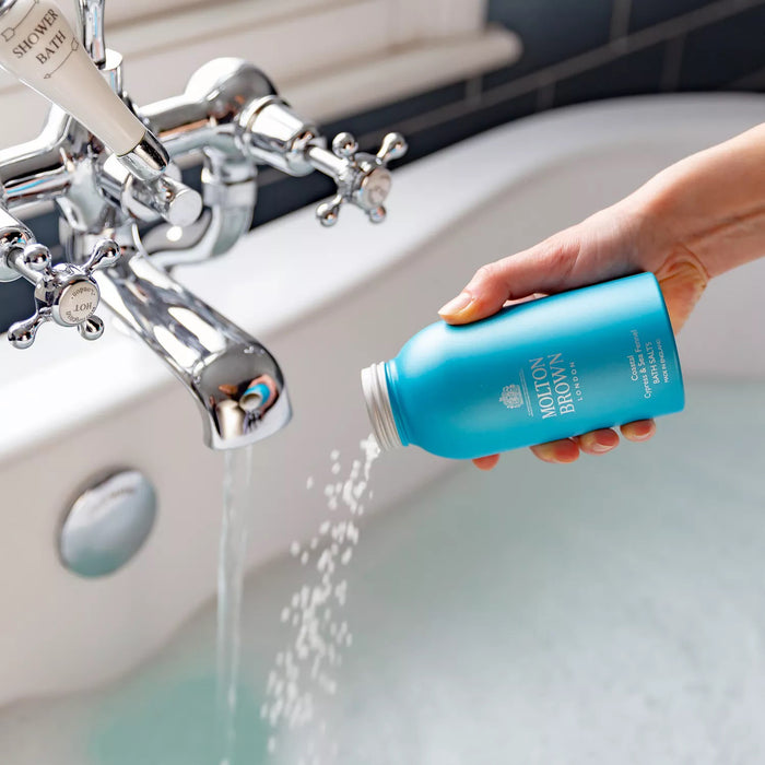 A hand pours MOLTON BROWN - COASTAL CYPRESS BATH SALTS, known for their marine notes, from a blue bottle into a bathtub while water streams in from a chrome faucet.