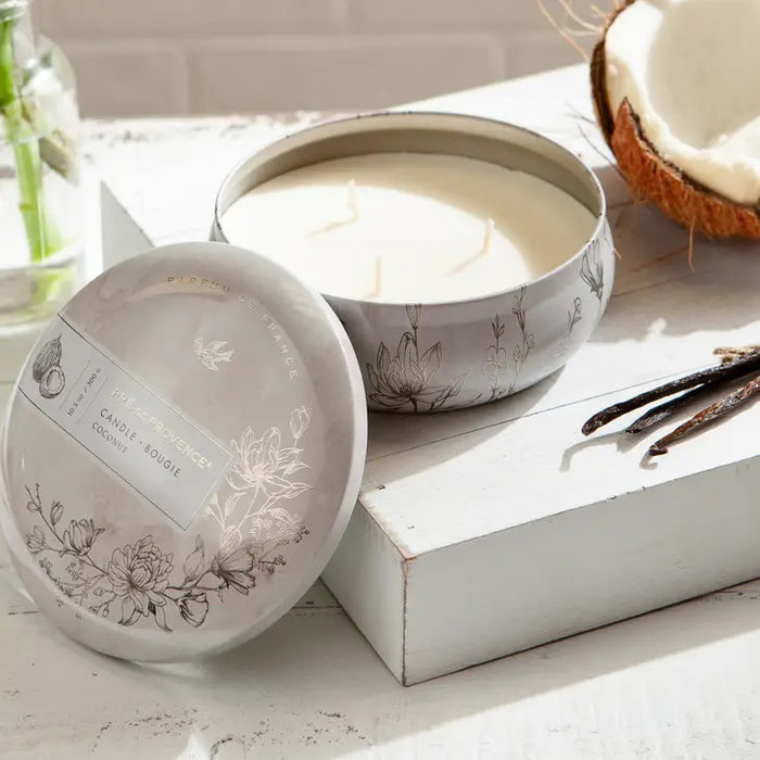 White round tin labeled “3 WICK CANDLE - COCONUT” by EUROPEAN SOAPS, LLC features floral art, coconut and bird motifs, and the phrase “Parfum de France.” Elegant minimalist lid holds a milky-scented candle for a refined home atmosphere.