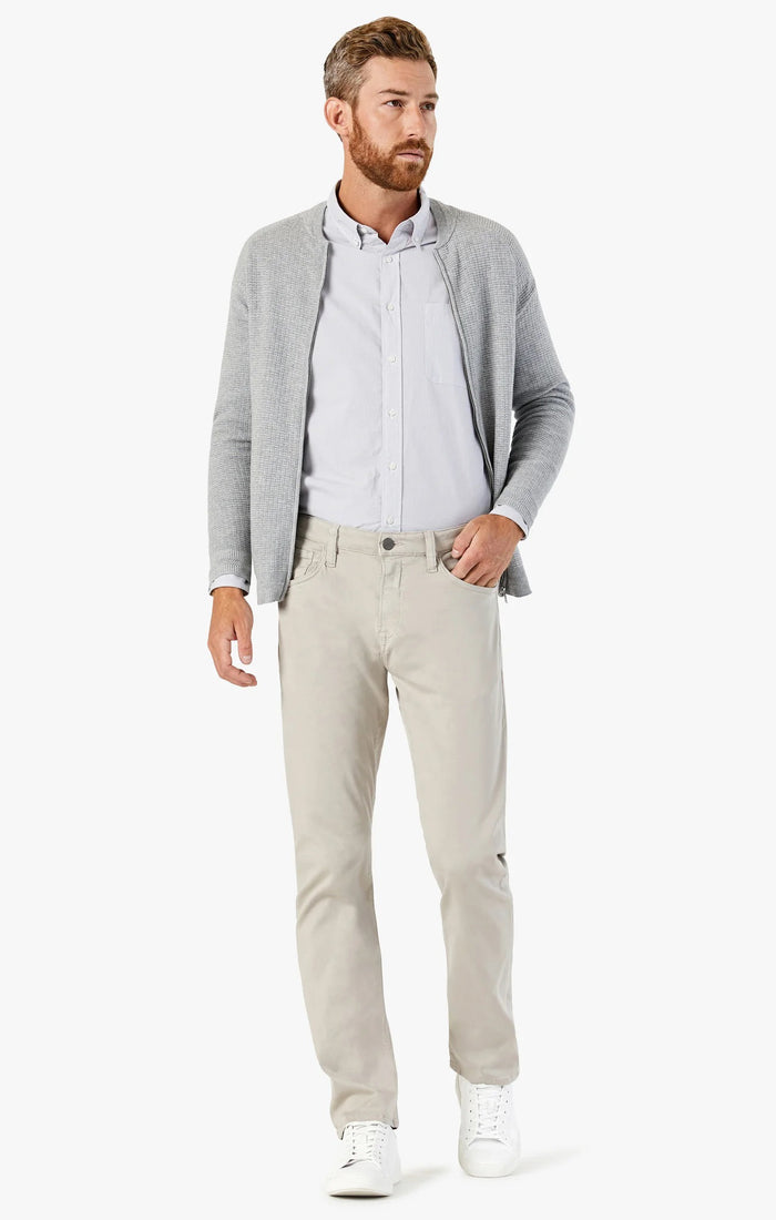 A bearded man stands facing forward with one hand in his pocket, wearing a light gray cardigan over a white button-up shirt, 34 HERITAGE - COURAGE STRAIGHT LEG PANTS IN DAWN TWILL (beige), and white sneakers against a plain white background.