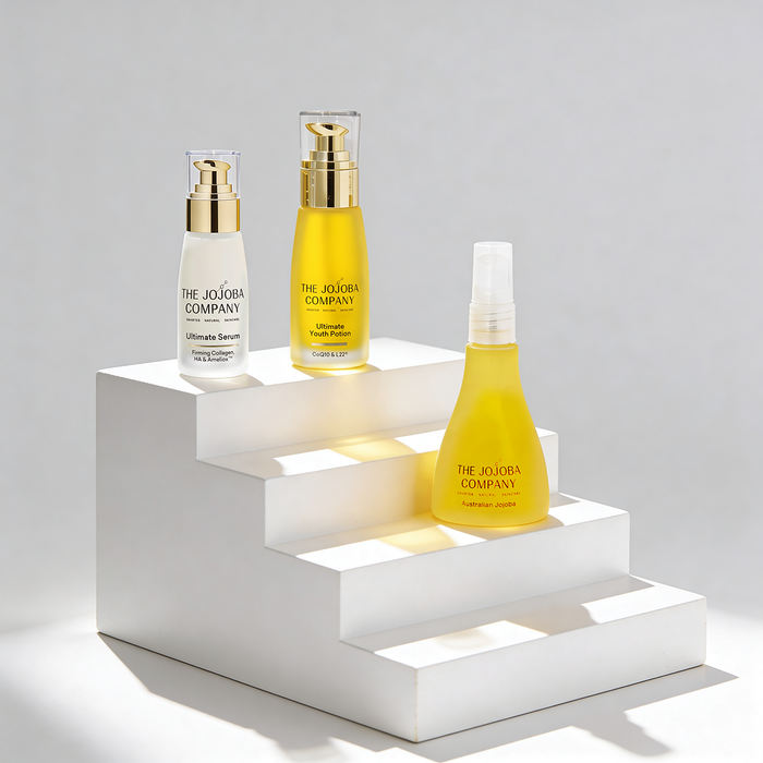 Three bottles of The Jojoba Company products on a white geometric shelf against a light gray background.