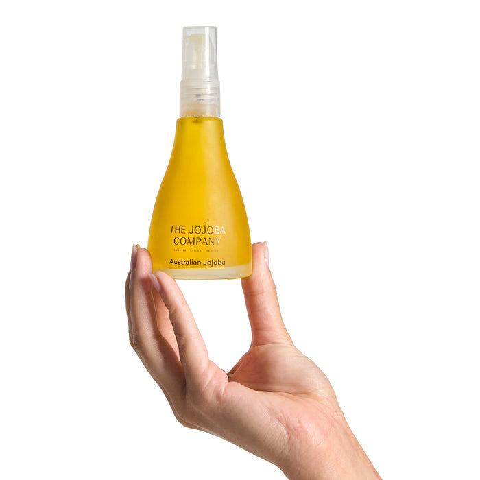 Hand holding a yellow bottle labeled 'The Jojoba Company' on a white background