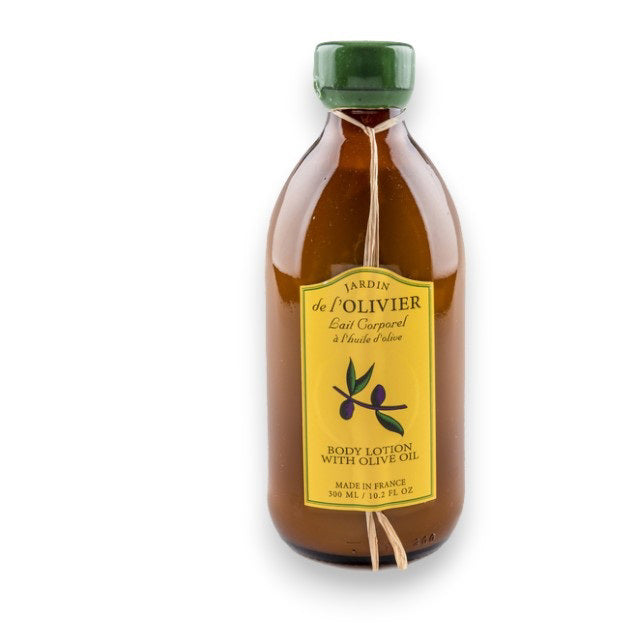 A brown glass bottle with a yellow label reading "BAUDELAIRE - JARDIN DE L'OLIVIER BODY LOTION," featuring an olive branch illustration and raffia bow. This natural lotion by BAUDELAIRE is ideal for daily unisex use.