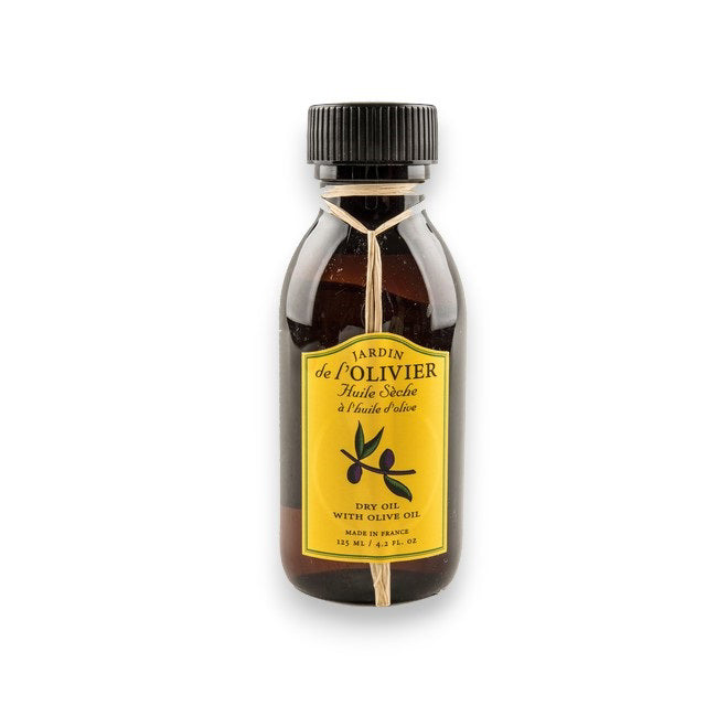 A brown bottle with a black cap, labeled "BAUDELAIRE - JARDIN DE L'OLIVIER DRY OIL." The yellow label features black text, an olive branch illustration, and notes the natural olive oil scent. A thin string is tied around the neck.