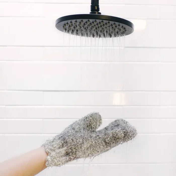 A hand wears the BAUDELAIRE - SISAL & HORSEHAIR BATH GLOVE under running water from a round black showerhead, set against a white tiled wall.
