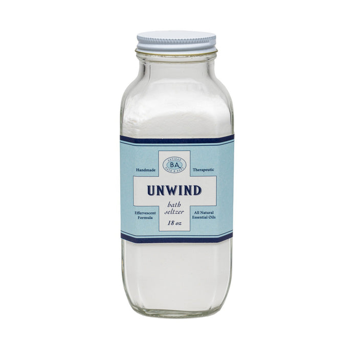 A clear glass jar of BAUDELAIRE - UNWIND BATH SELTZER 18 OZ, featuring a blue and white label noting handmade, therapeutic, effervescent soaks with all-natural essential oils.