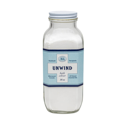 A clear glass jar of BAUDELAIRE - UNWIND BATH SELTZER 18 OZ, featuring a blue and white label noting handmade, therapeutic, effervescent soaks with all-natural essential oils.