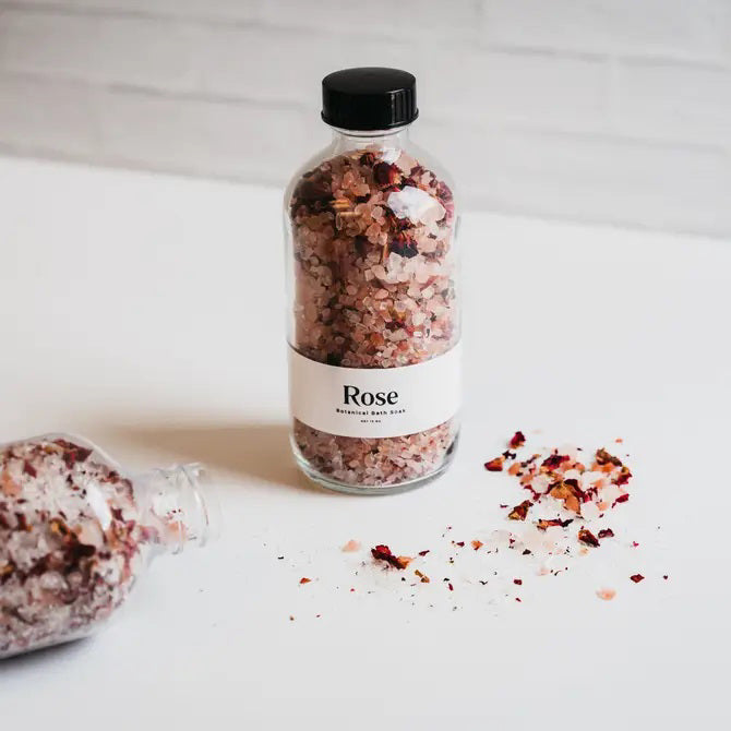 A clear bottle from GIFTEN MARKET holds pink and white BOTANICAL BATH SOAK - ROSE with dried rose petals. The label displays "Rose Botanical Bath Soak" on a white background.