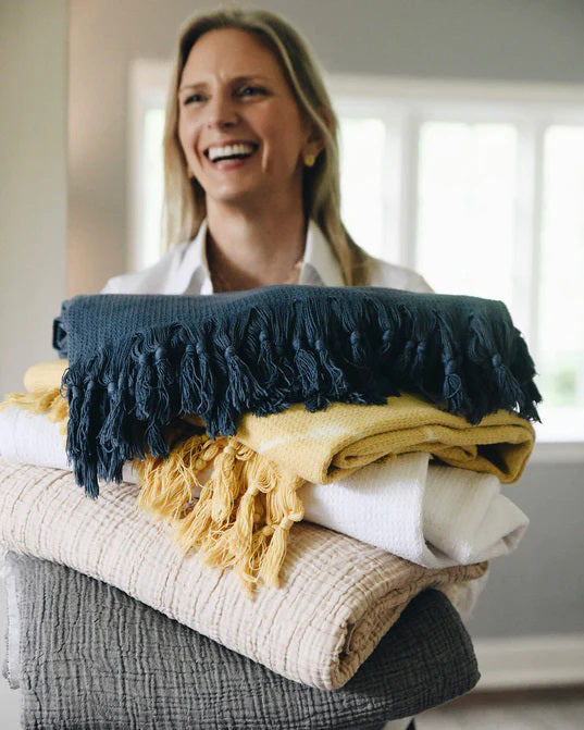 The MERSEA CLOUD GAUZE BLANKET, shown folded on a light background, is a soft beige cotton throw with a textured surface and fringed edges.