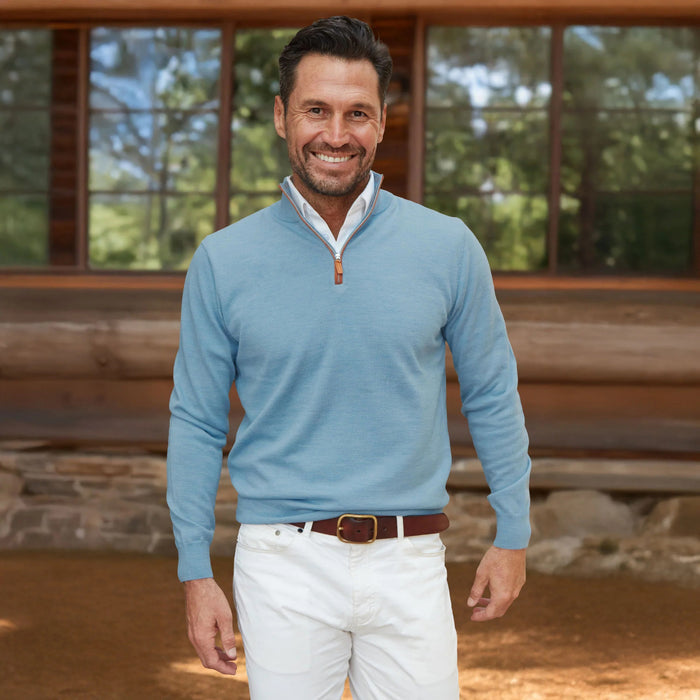 A man with dark hair and a beard smiles outside a wooden building, wearing COLLARS AND CO’s Blue Melange with Brown Suede Trim Cashmere Touch Australian Merino Quarter Zip Sweater, paired with white pants and a brown belt.