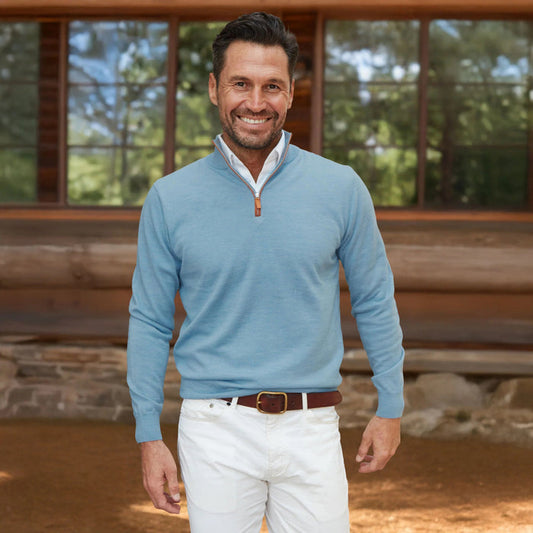 A man with dark hair and a beard smiles outside a wooden building, wearing COLLARS AND CO’s Blue Melange with Brown Suede Trim Cashmere Touch Australian Merino Quarter Zip Sweater, paired with white pants and a brown belt.