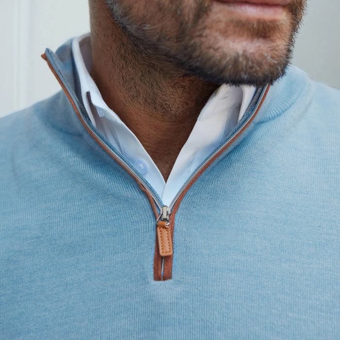 A man with dark hair and a beard smiles outside a wooden building, wearing COLLARS AND CO’s Blue Melange with Brown Suede Trim Cashmere Touch Australian Merino Quarter Zip Sweater, paired with white pants and a brown belt.