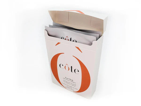 A white package with orange accents, partially open to show multiple sheet masks inside. The label reads "COTE," highlighting plant-based collagen for age-defying hydration. Product: COTE - FOOT MASK WITH COLLAGEN 3 PACK.