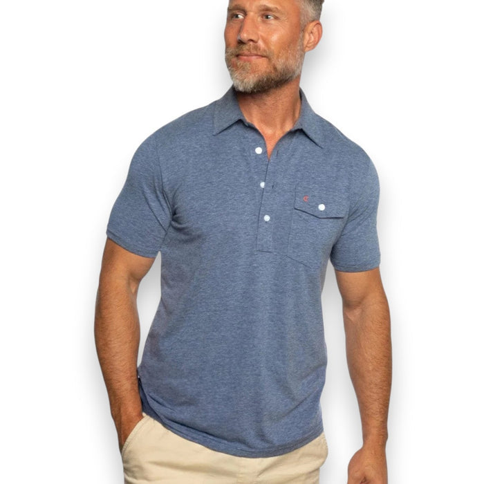 A man with short gray hair and a beard wears the CRIQUET Performance Players Shirt in Midnight, a moisture-wicking classic fit polo with a chest pocket, paired with light pants. He stands with one hand in his pocket against a white background.