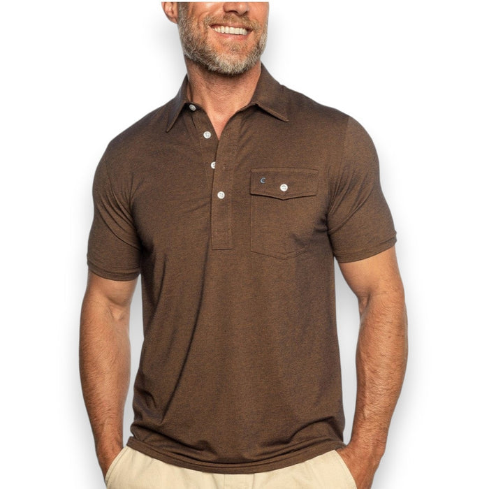 A man stands smiling with one hand in his pocket, wearing the CRIQUET Performance Players Shirt in Woodsmoke—a short-sleeve, brown, moisture-wicking polo with a chest pocket and white buttons—paired with light-colored pants against a white background.