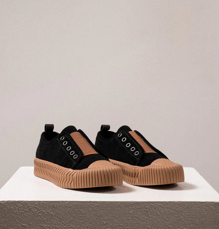 The DANIELLA LEHAVI - OSAKA SNEAKER in black, designed for daily wear, features thick tan rubber soles and a brown fabric strip, shown on a white textured pedestal with a neutral background.
