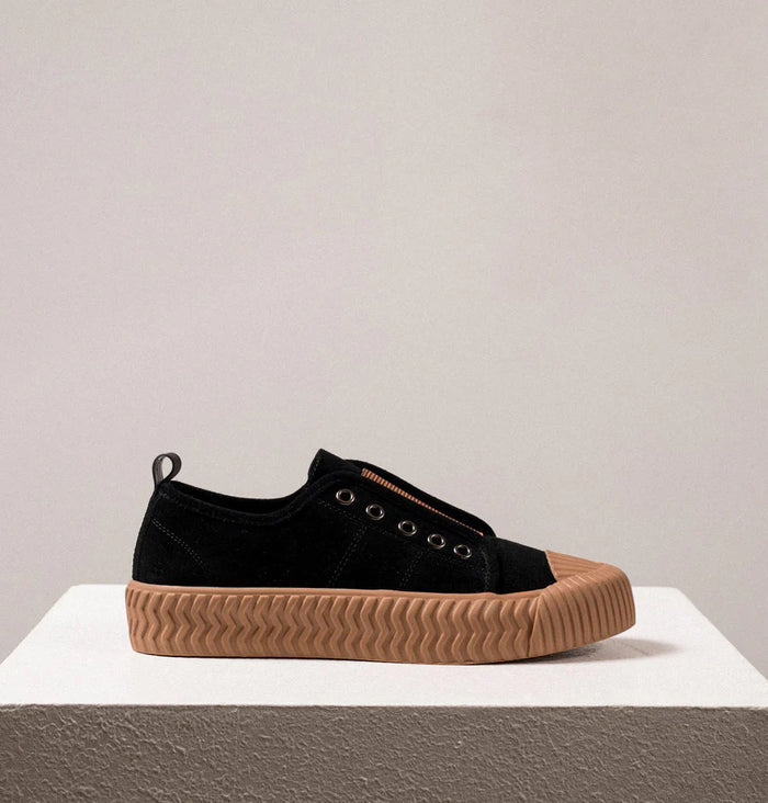 The DANIELLA LEHAVI - OSAKA SNEAKER in black, designed for daily wear, features thick tan rubber soles and a brown fabric strip, shown on a white textured pedestal with a neutral background.