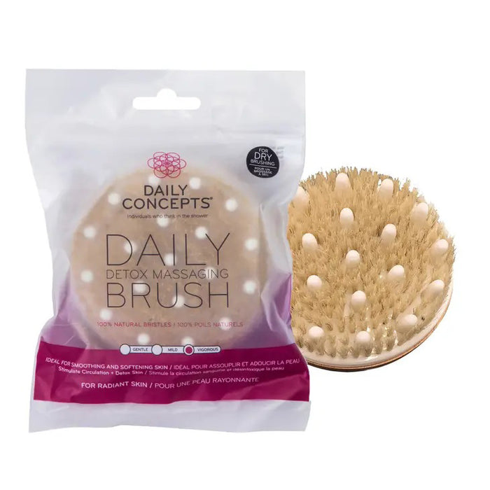 The DAILY CONCEPTS DETOX MASSAGE BRUSH, featuring natural bristles, is shown next to its clear packaging that highlights benefits like dry brushing for smoother, more radiant skin and helping reduce cellulite.