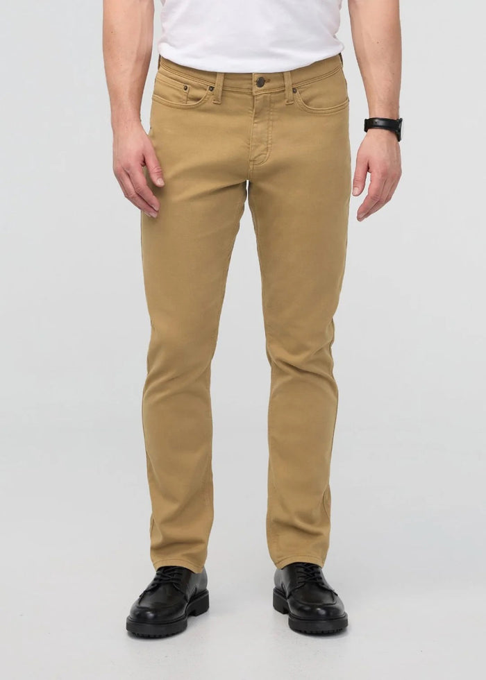 A man with short dark hair and a trimmed beard stands against a white background, wearing a white t-shirt, white sneakers with black details, and DUER's DU/ER NO SWEAT RELAXED TAPER PANT in an athletic fit. One hand is in his pocket.