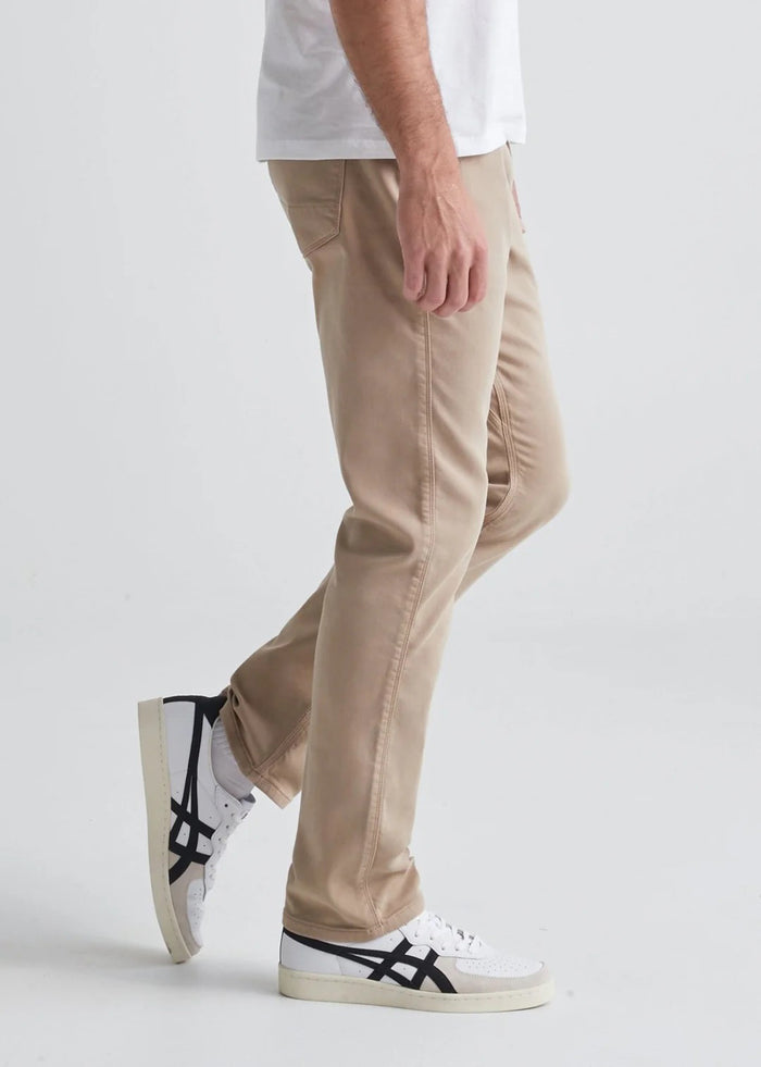 A man with short dark hair and a trimmed beard stands against a white background, wearing a white t-shirt, white sneakers with black details, and DUER's DU/ER NO SWEAT RELAXED TAPER PANT in an athletic fit. One hand is in his pocket.