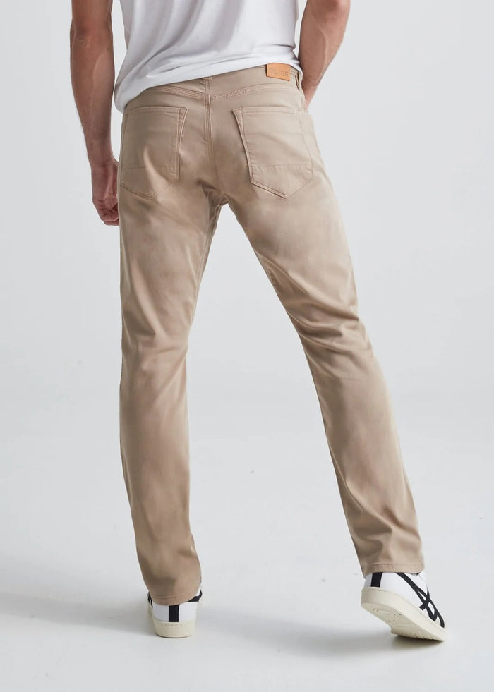 A man with short dark hair and a trimmed beard stands against a white background, wearing a white t-shirt, white sneakers with black details, and DUER's DU/ER NO SWEAT RELAXED TAPER PANT in an athletic fit. One hand is in his pocket.