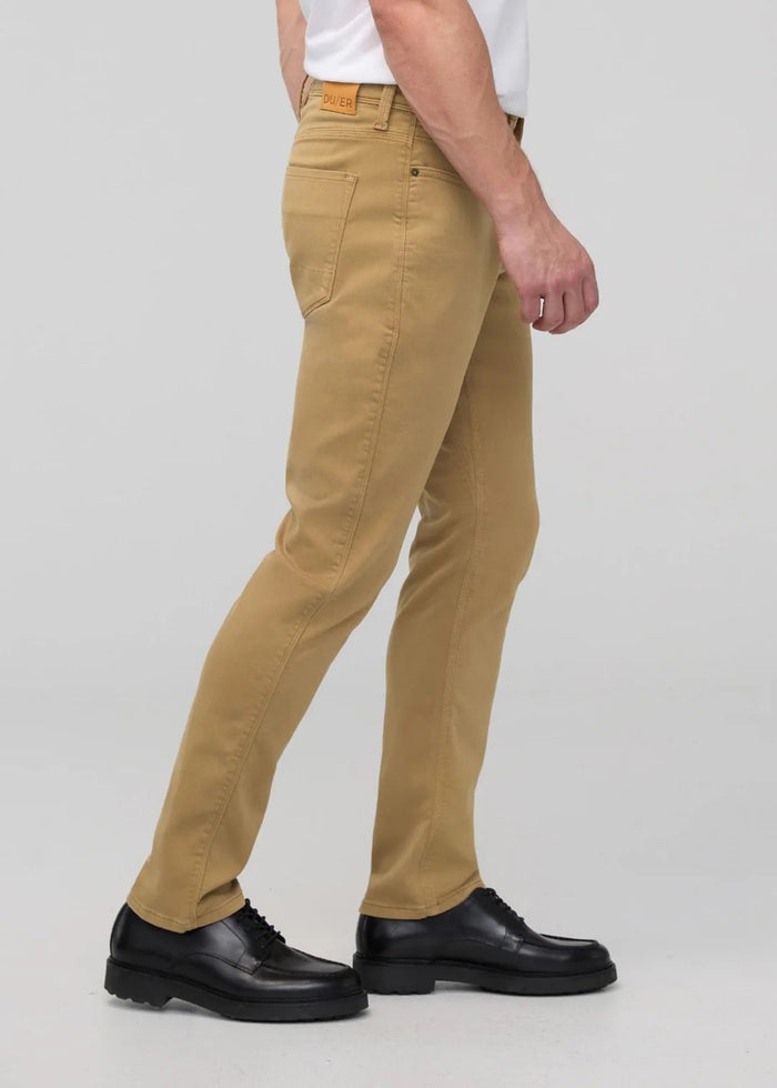 A man with short dark hair and a trimmed beard stands against a white background, wearing a white t-shirt, white sneakers with black details, and DUER's DU/ER NO SWEAT RELAXED TAPER PANT in an athletic fit. One hand is in his pocket.