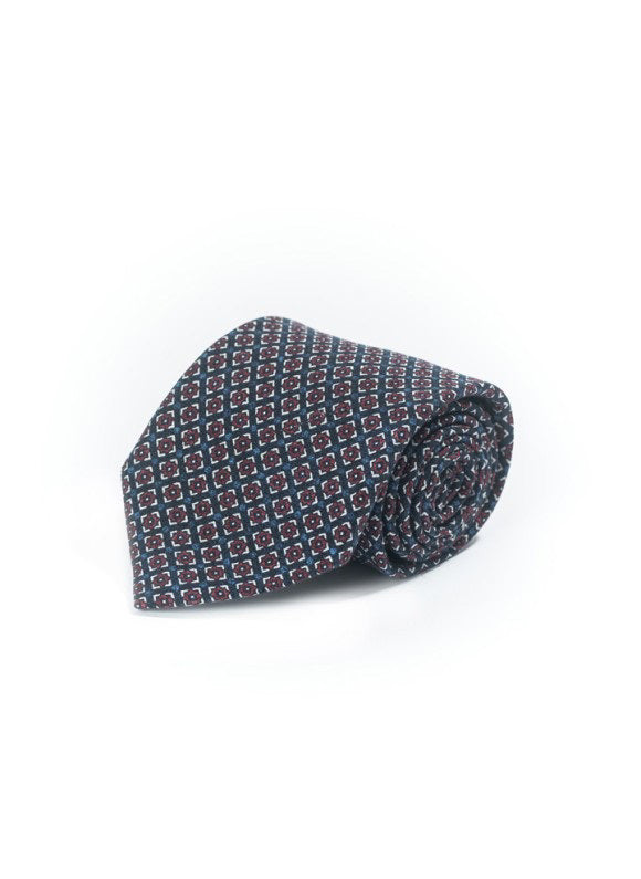 The EDWARD ARMAH - NAVY NEAT SILK TIE, featuring a repeating geometric pattern in blue, red, and white, is rolled up and displayed against a plain white background.