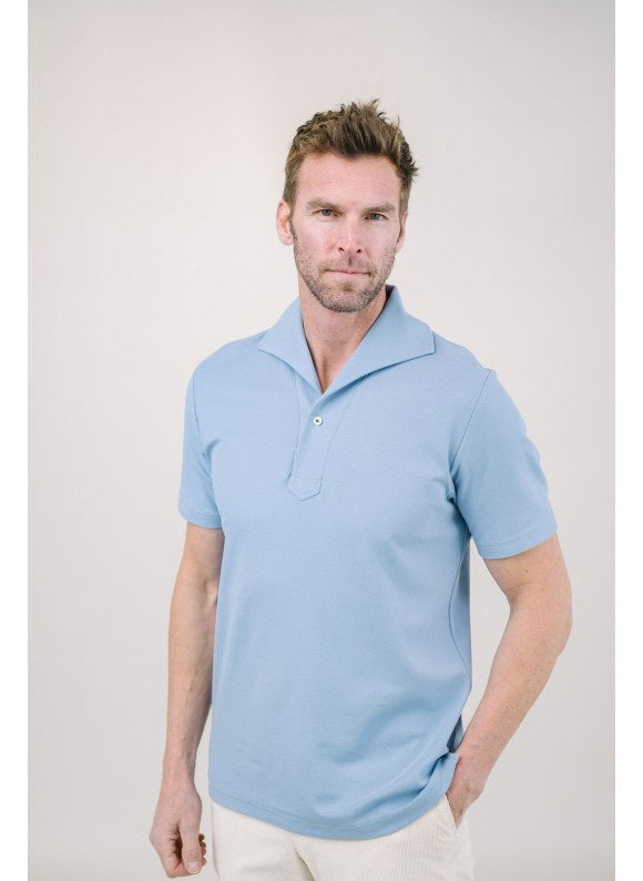 A man with light brown hair and a short beard wears the EDWARD ARMAH Pima Cotton Tailored Polo Shirt in dark blue and jeans, standing against a plain light background with a relaxed expression and one hand in his pocket.