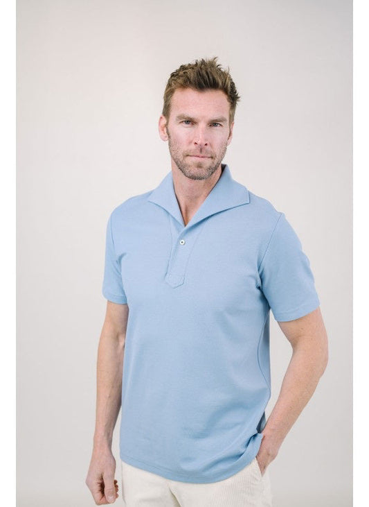 A man with light brown hair and a short beard wears the EDWARD ARMAH Pima Cotton Tailored Polo Shirt in dark blue and jeans, standing against a plain light background with a relaxed expression and one hand in his pocket.