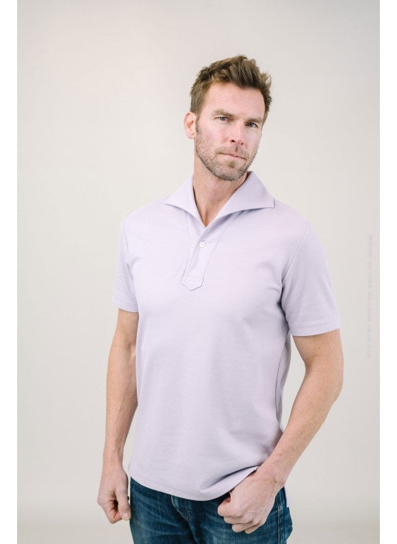 A man with light brown hair and a short beard wears the EDWARD ARMAH Pima Cotton Tailored Polo Shirt in dark blue and jeans, standing against a plain light background with a relaxed expression and one hand in his pocket.