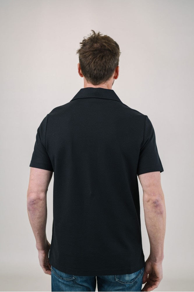 A man with light brown hair and a short beard wears the EDWARD ARMAH Pima Cotton Tailored Polo Shirt in dark blue and jeans, standing against a plain light background with a relaxed expression and one hand in his pocket.