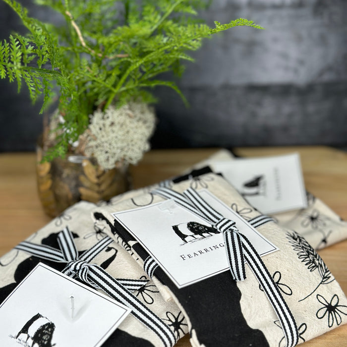 The CALHOUN & CO. FEARRINGTON SIGNATURE COLLECTION - BESPOKE BELTIE BARNYARD TEA TOWEL features black, hand-drawn belted cows, goats, and chickens with florals and a wooden fence, capturing the charm of Fearrington Village.