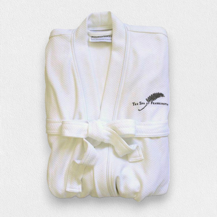 A FEARRINGTON SIGNATURE COLLECTION - FEARRINGTON SPA ROBE by BOCA, white with a belt and "The Spa at Fearrington" plus fern logo embroidered on the chest, is neatly folded on a light textured surface.