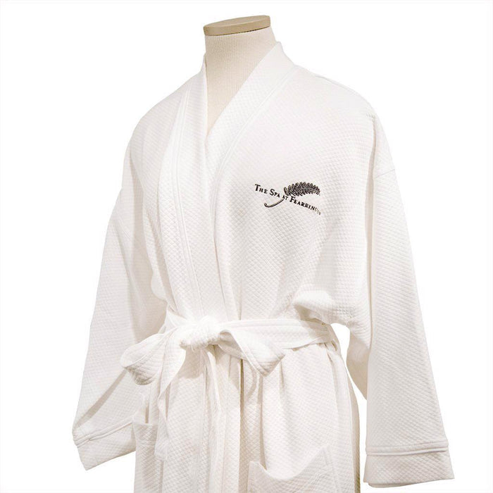 A FEARRINGTON SIGNATURE COLLECTION - FEARRINGTON SPA ROBE by BOCA, white with a belt and "The Spa at Fearrington" plus fern logo embroidered on the chest, is neatly folded on a light textured surface.
