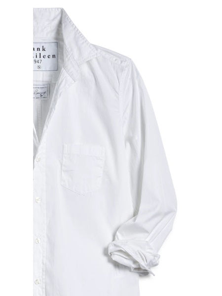 A man with short curly hair wears the FRANK & EILEEN LUKE Men's Button Up Shirt in Classic White Poplin (sleeves rolled), paired with light blue ripped jeans and a watch, smiling slightly against a plain background.