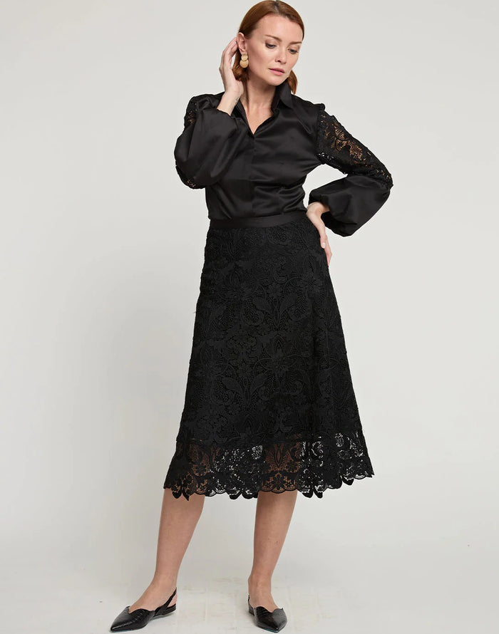 A woman stands against a plain background, wearing a black satin blouse with lace sleeves, the HINSON WU - GLORIA LACE SKIRT by HINSON WU, and chic pointed-toe black shoes, with one hand raised near her ear.
