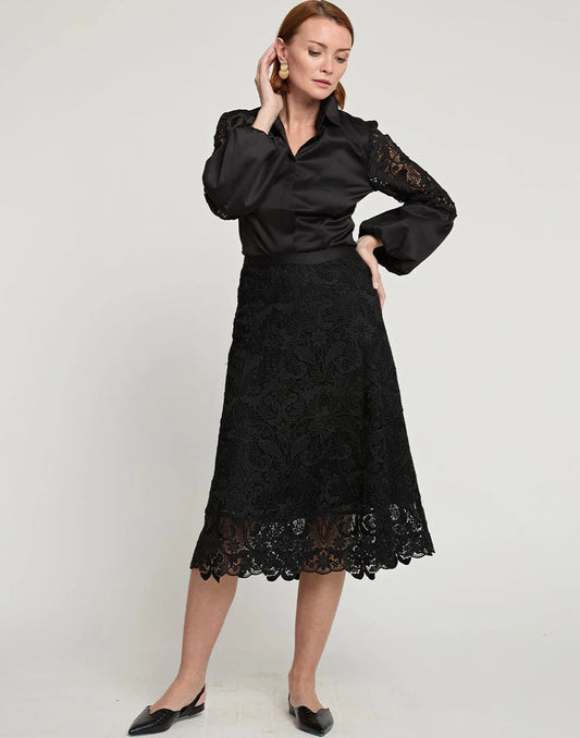 A woman stands against a plain background, wearing a black satin blouse with lace sleeves, the HINSON WU - GLORIA LACE SKIRT by HINSON WU, and chic pointed-toe black shoes, with one hand raised near her ear.