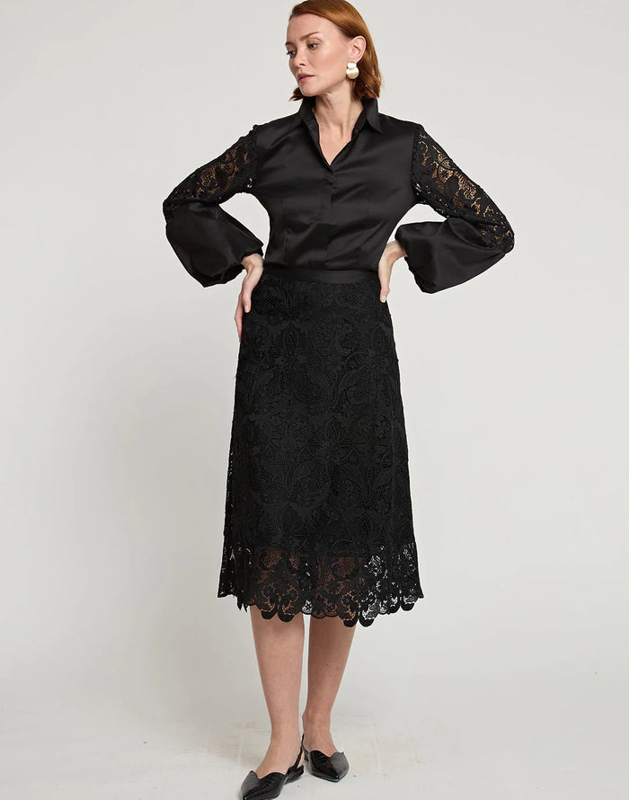 A woman stands against a plain background, wearing a black satin blouse with lace sleeves, the HINSON WU - GLORIA LACE SKIRT by HINSON WU, and chic pointed-toe black shoes, with one hand raised near her ear.