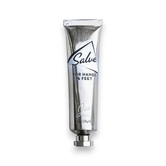 A metallic, recyclable tube labeled “JAO BRAND - SALVE FOR HANDS AND FEET” from JAO LIMITED stands upright on a white background with a silver cap and appears partially squeezed, suggesting its nourishing blend of essential oils.