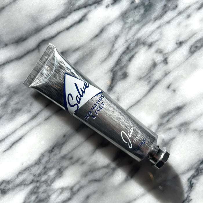 A metallic, recyclable tube labeled “JAO BRAND - SALVE FOR HANDS AND FEET” from JAO LIMITED stands upright on a white background with a silver cap and appears partially squeezed, suggesting its nourishing blend of essential oils.
