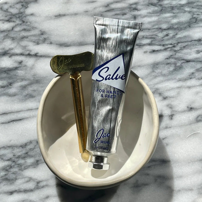 A metallic, recyclable tube labeled “JAO BRAND - SALVE FOR HANDS AND FEET” from JAO LIMITED stands upright on a white background with a silver cap and appears partially squeezed, suggesting its nourishing blend of essential oils.