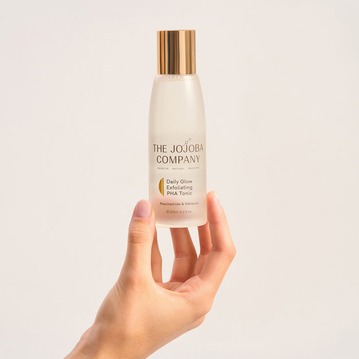 Hand holding a bottle of 'The Jojoba Company' skincare product against a light background