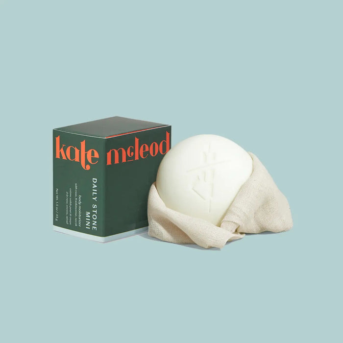 A round white KATE MCLEOD - DAILY STONE MINI LOTION BAR 1OZ, partially wrapped in beige cloth, rests beside its green box on a pale blue background. Crafted with natural essential oils for rich hydration.