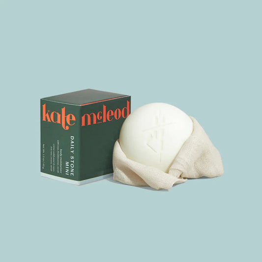 A round white KATE MCLEOD - DAILY STONE MINI LOTION BAR 1OZ, partially wrapped in beige cloth, rests beside its green box on a pale blue background. Crafted with natural essential oils for rich hydration.